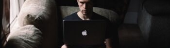 Man sitting on a sofa in a gloomy room looking at his laptop screen with a concerned expression.