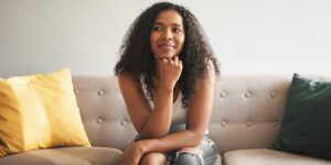 Young black woman sitting on a sofa looking thoughtful and smiling slightly